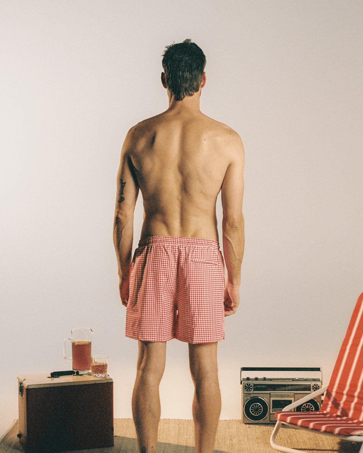 A man stands with his back to the camera wearing red and white gingham checkered swim trunks, set against a minimalist studio background with vintage props.