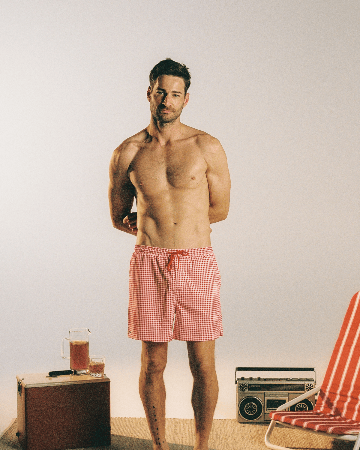 A shirtless man stands barefoot in a studio setting wearing red and white gingham check swim trunks. A vintage suitcase with a drink, a retro boombox, and part of a striped beach chair are placed on the floor around him against a plain light background.