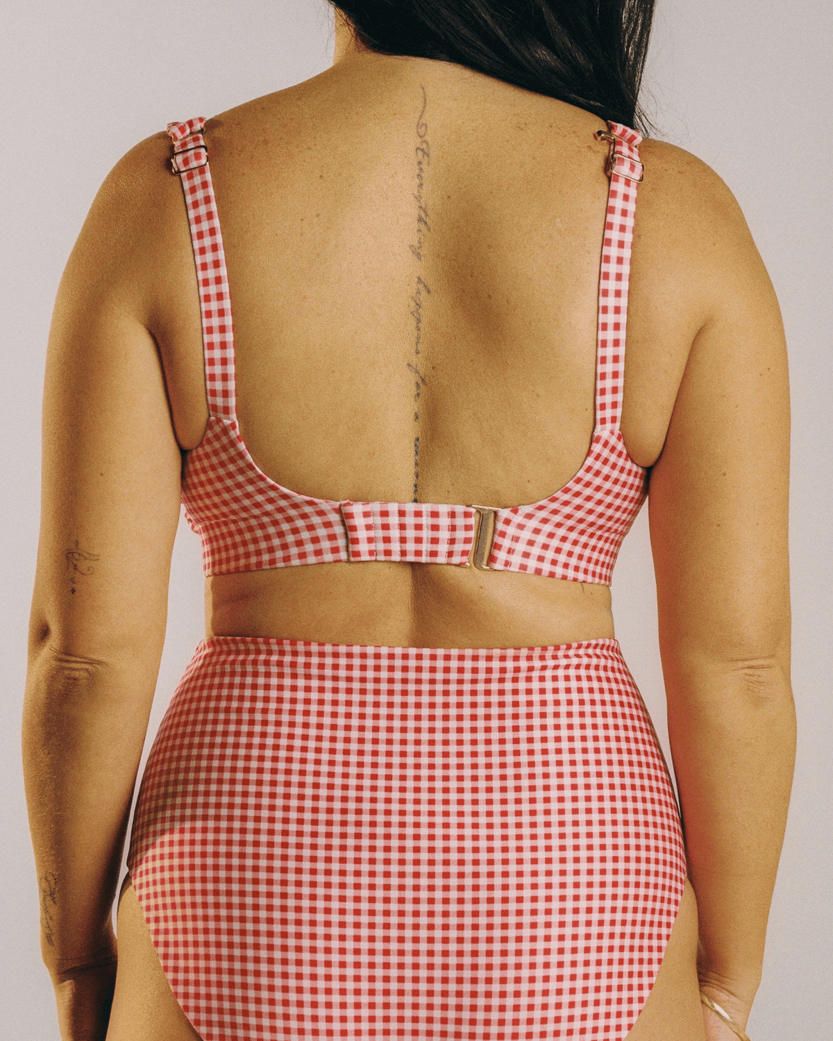 A woman shown from the back wearing a red and white gingham two-piece swimsuit with a cutout waist and adjustable straps, against a plain light white studio background.