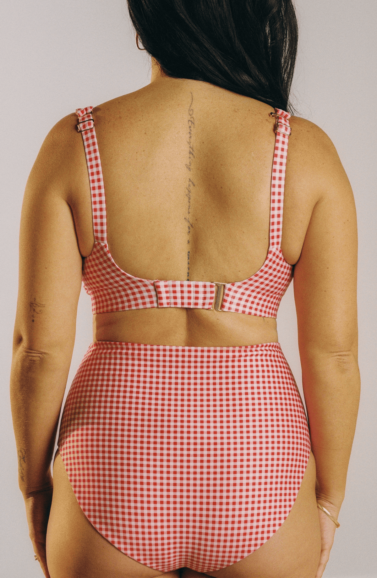 A woman shown from the back wearing a red and white gingham two-piece swimsuit with a cutout waist and adjustable straps, against a plain light white studio background.