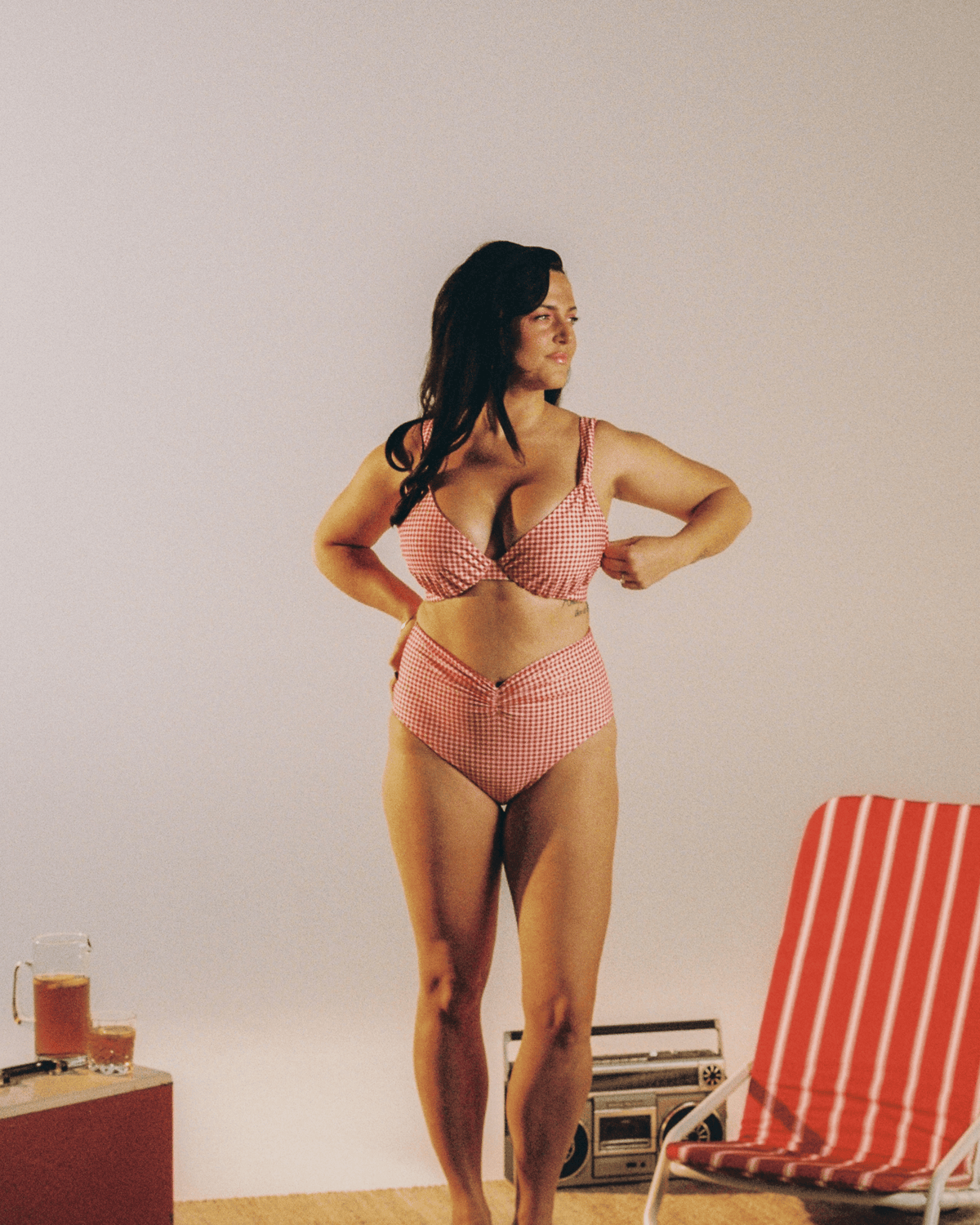 A woman stands barefoot indoors wearing a red and white gingham two-piece bikini. A red-and-white striped lounge chair and a small table with drinks are visible against a plain light wall.