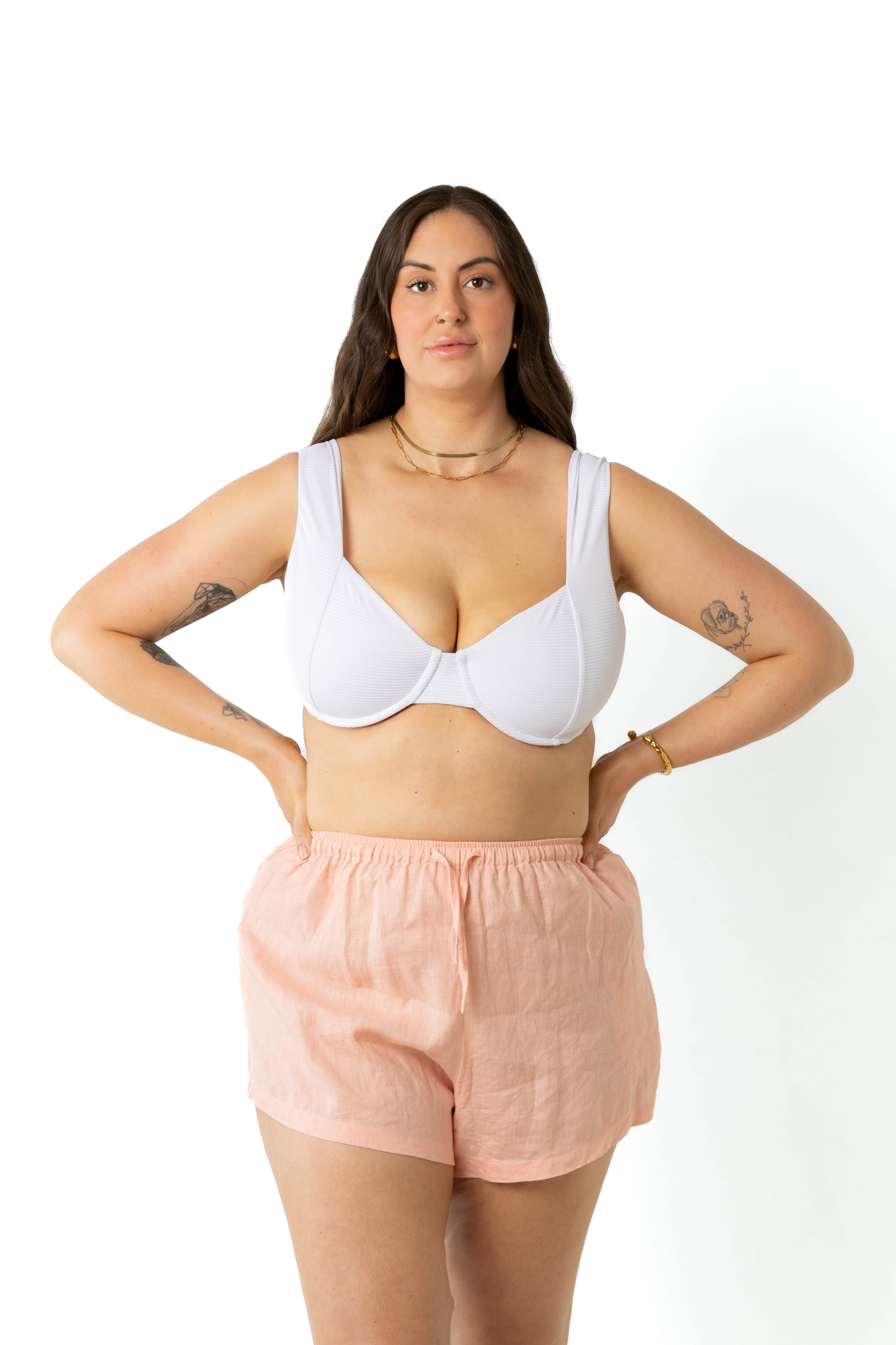 model in a photography studio facing camera, wearing a white ribbed underwire bikini top with peach colour linen shorts with her hands placed on her hips.