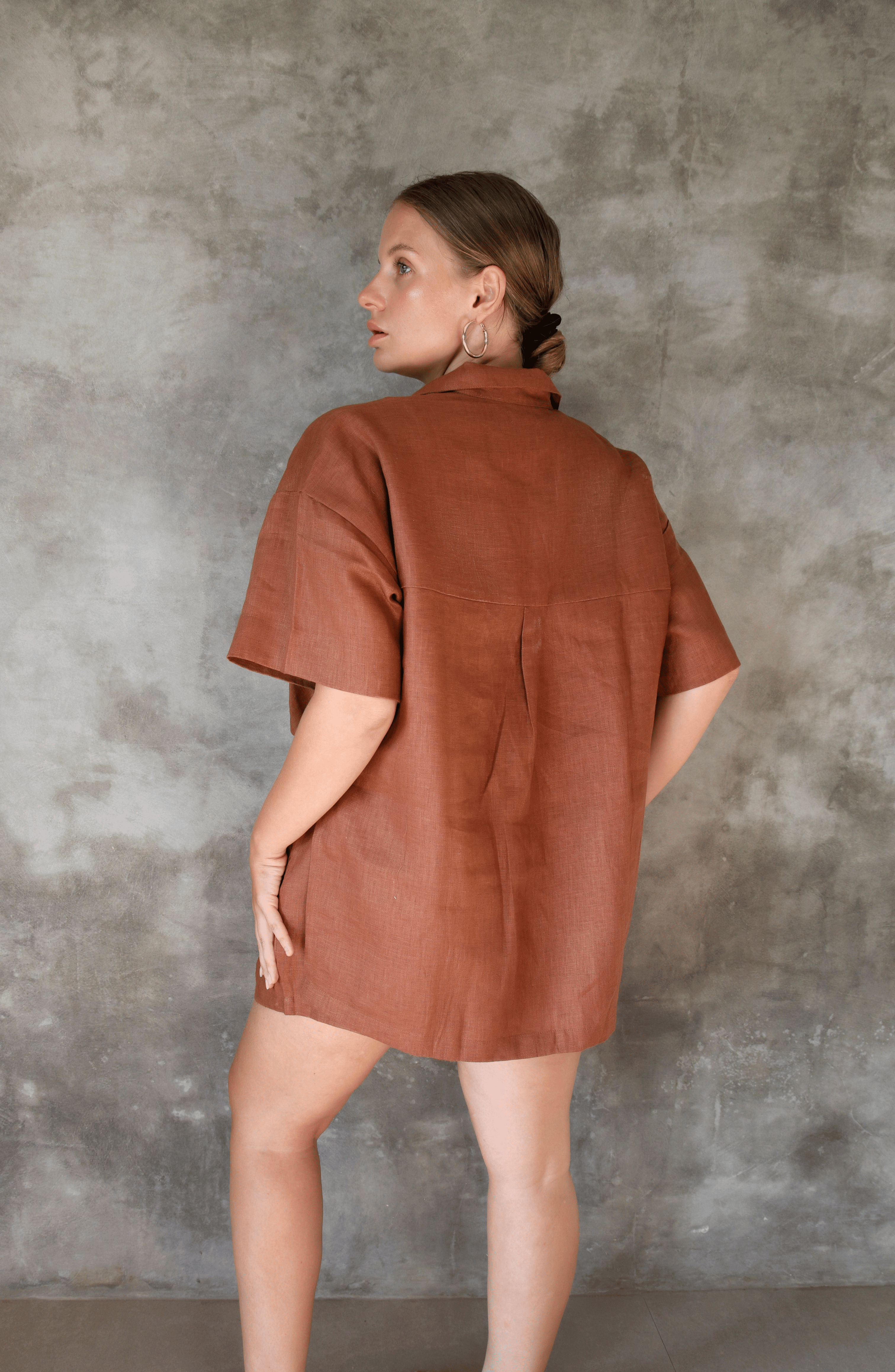 Model poses for photo in Pinksalt Swimwear klum espresso brown Linen shirt and shorts set. This is a back view, with her hands down by her side.
