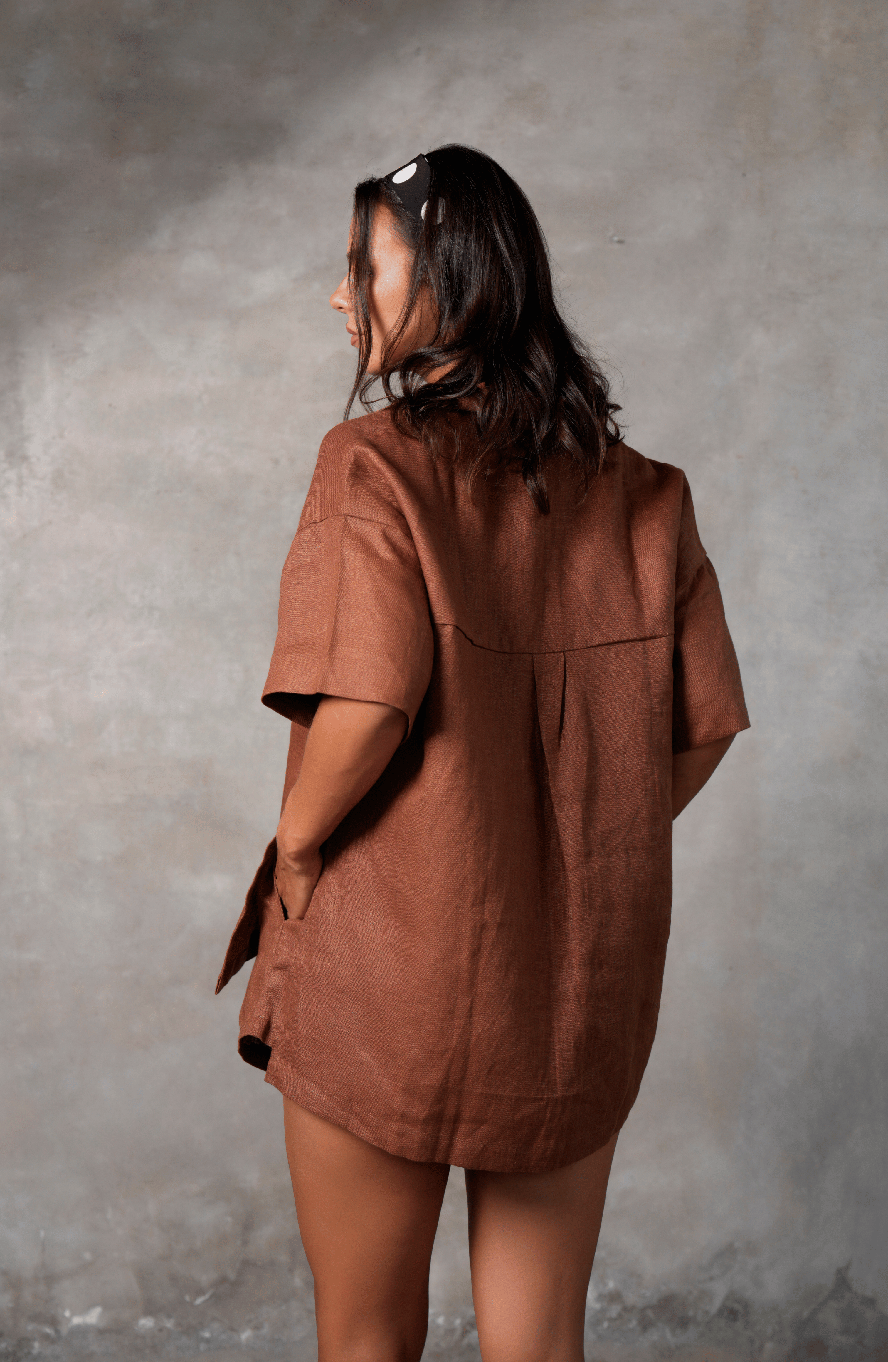 Model poses for photo in Pinksalt Swimwear klum espresso brown Linen shirt and shorts set. This is a back view, with her hands in the pocket of the linen shorts and the shirt untucked. Model is wearing polkadot head scarf.