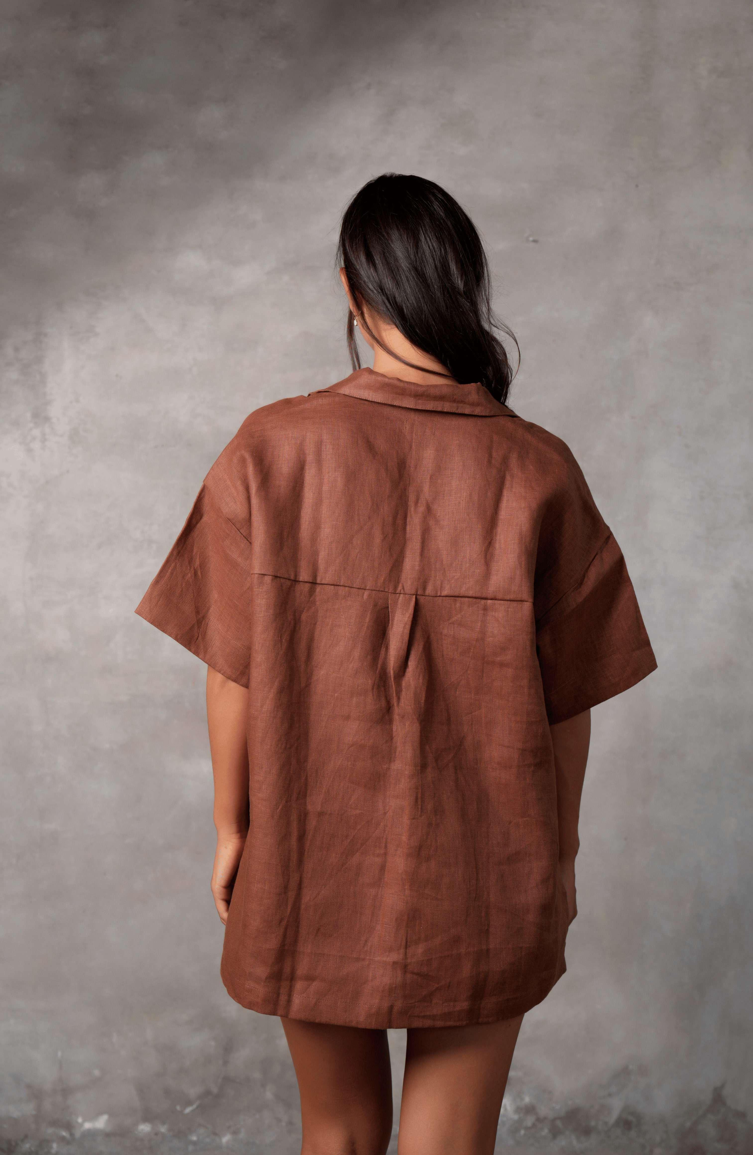 Model poses for photo in Pinksalt Swimwear klum espresso brown Linen shirt and shorts set. This is a back view, with her hands by her side. Model has hair to the side