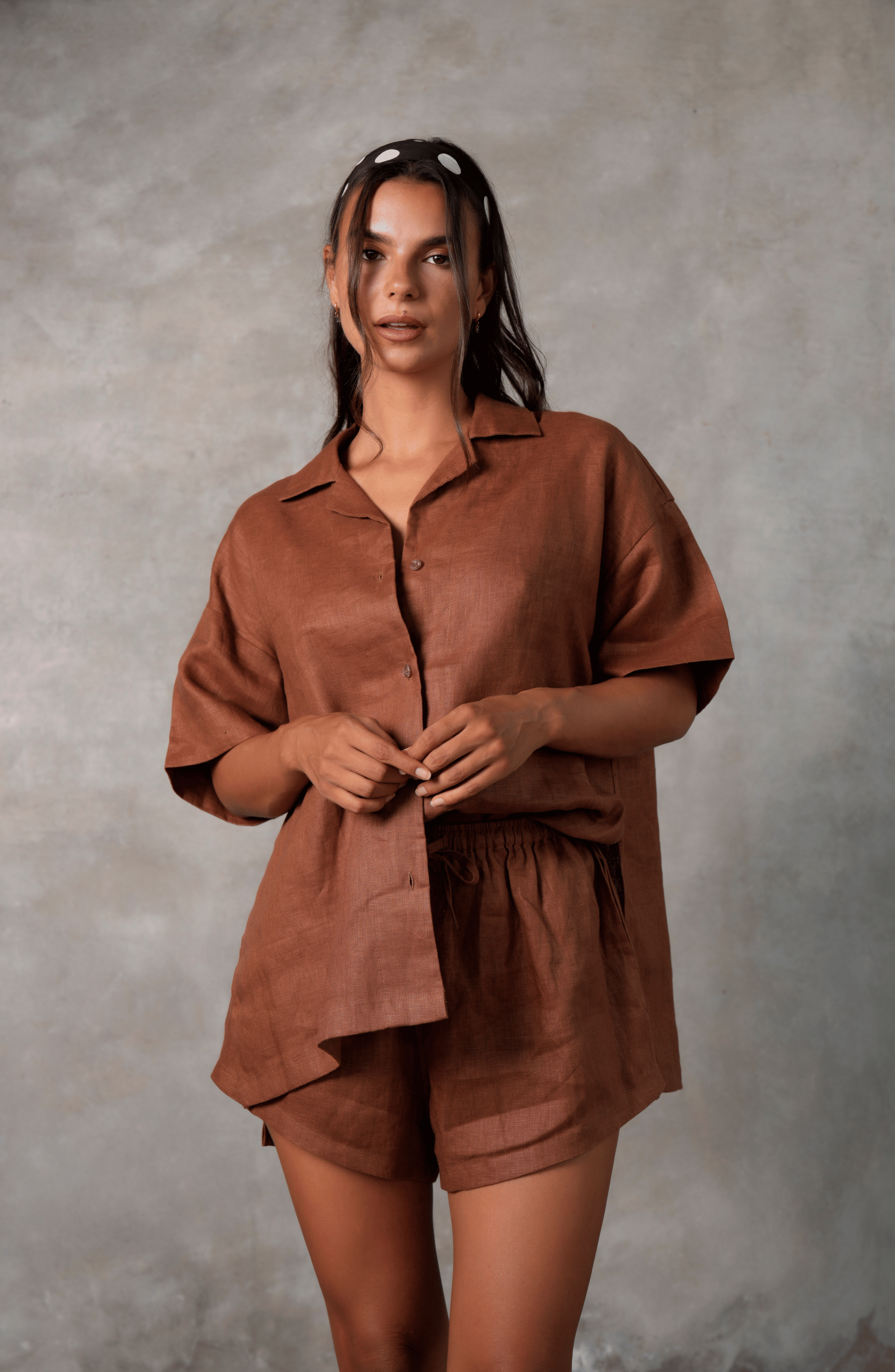 Model poses for photo in Pinksalt Swimwear klum espresso brown Linen shirt and shorts set. This is a front view, with her hands held in front of her, with one side of the linen shirt tucked into the drawstring linen shorts. Model is wearing polkadot head scarf.