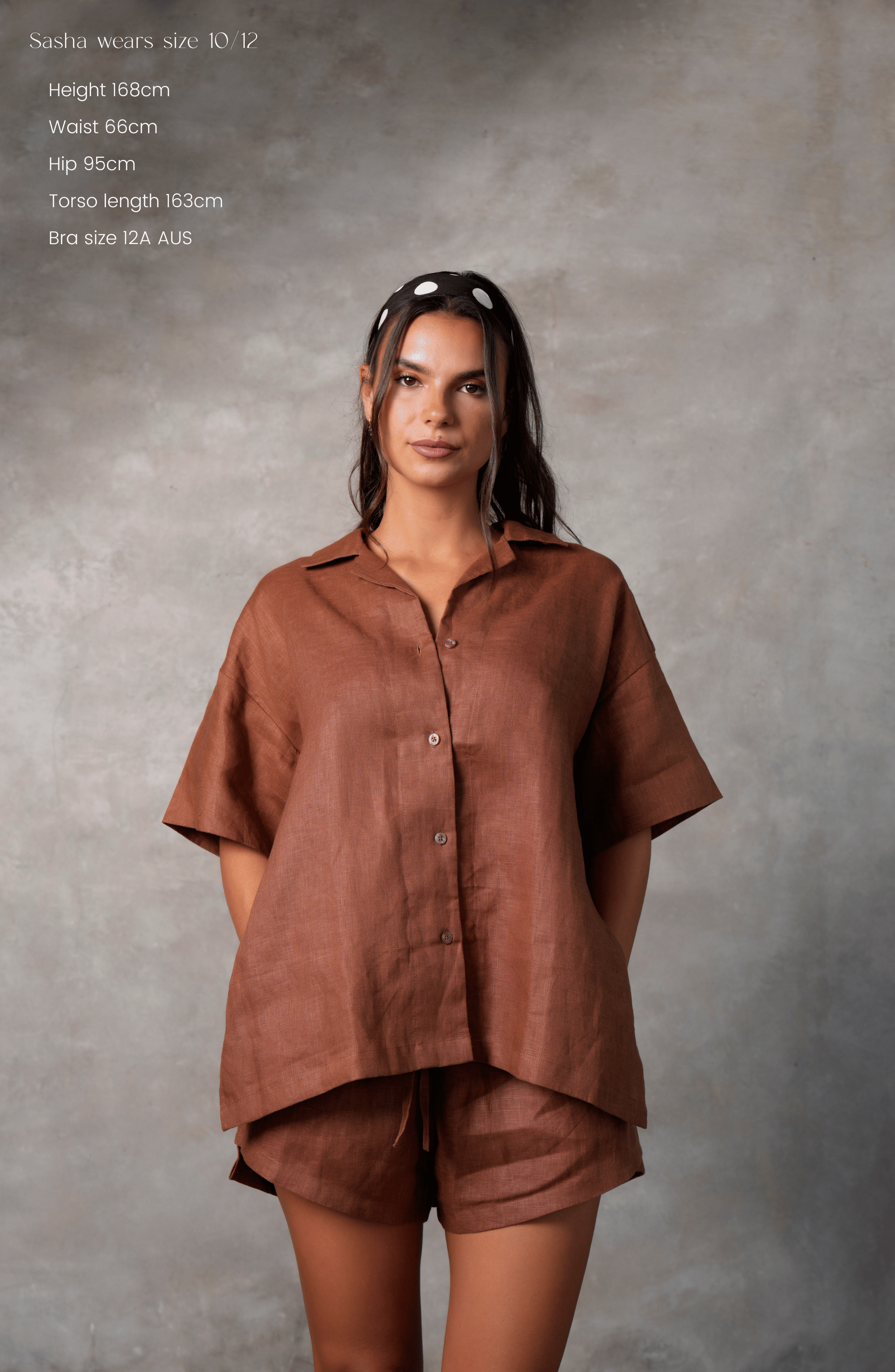 Model poses for photo in Pinksalt Swimwear klum espresso brown Linen shirt and shorts set. This is a front view, with her hands in the pocket of the linen shorts and the shirt untucked. Model is wearing polkadot head scarf.