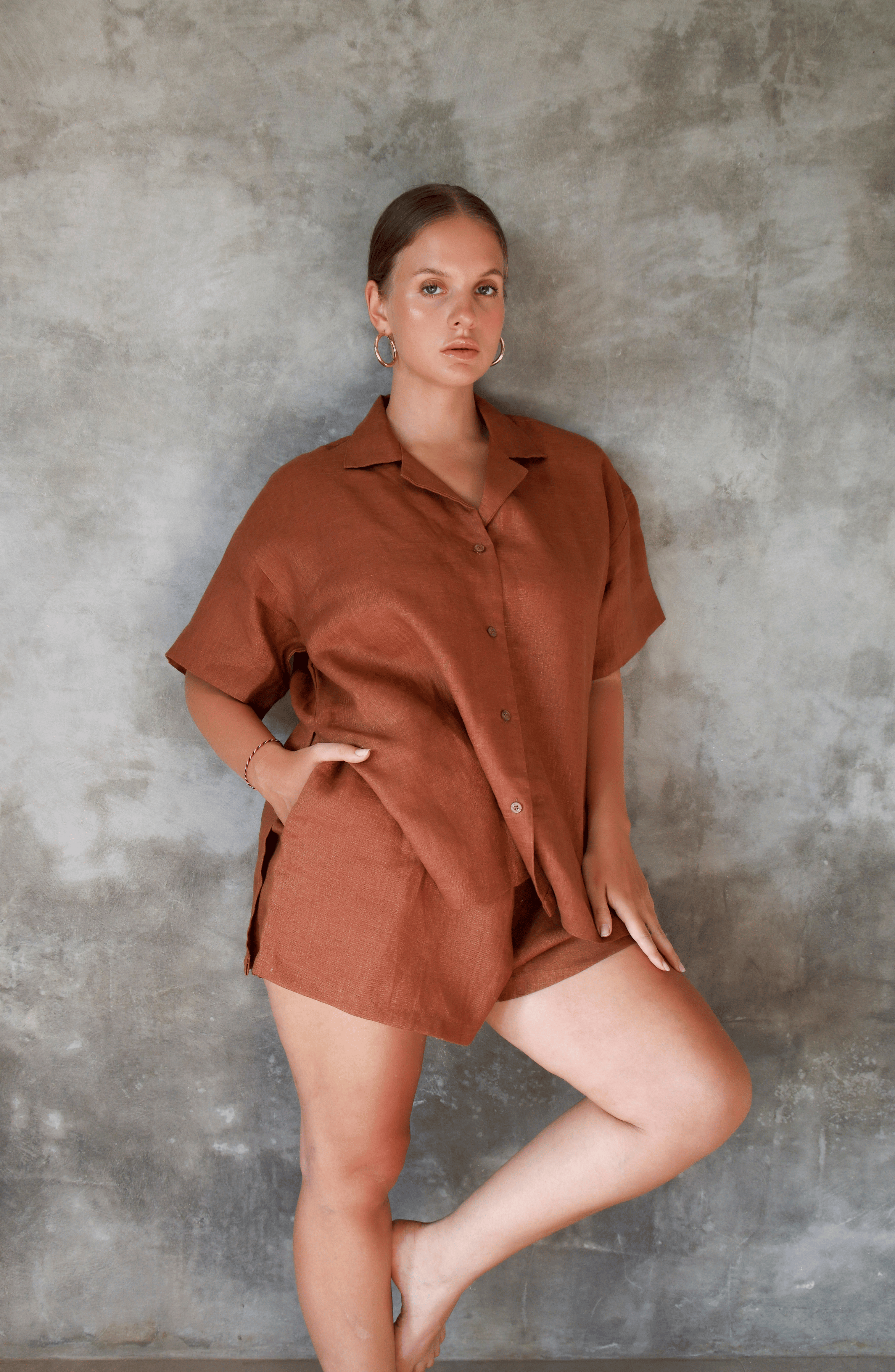 Model poses for photo in Pinksalt Swimwear klum espresso brown Linen shirt and shorts set. This is a front/side view, with her hand in the pocket of the drawstring linen shorts.