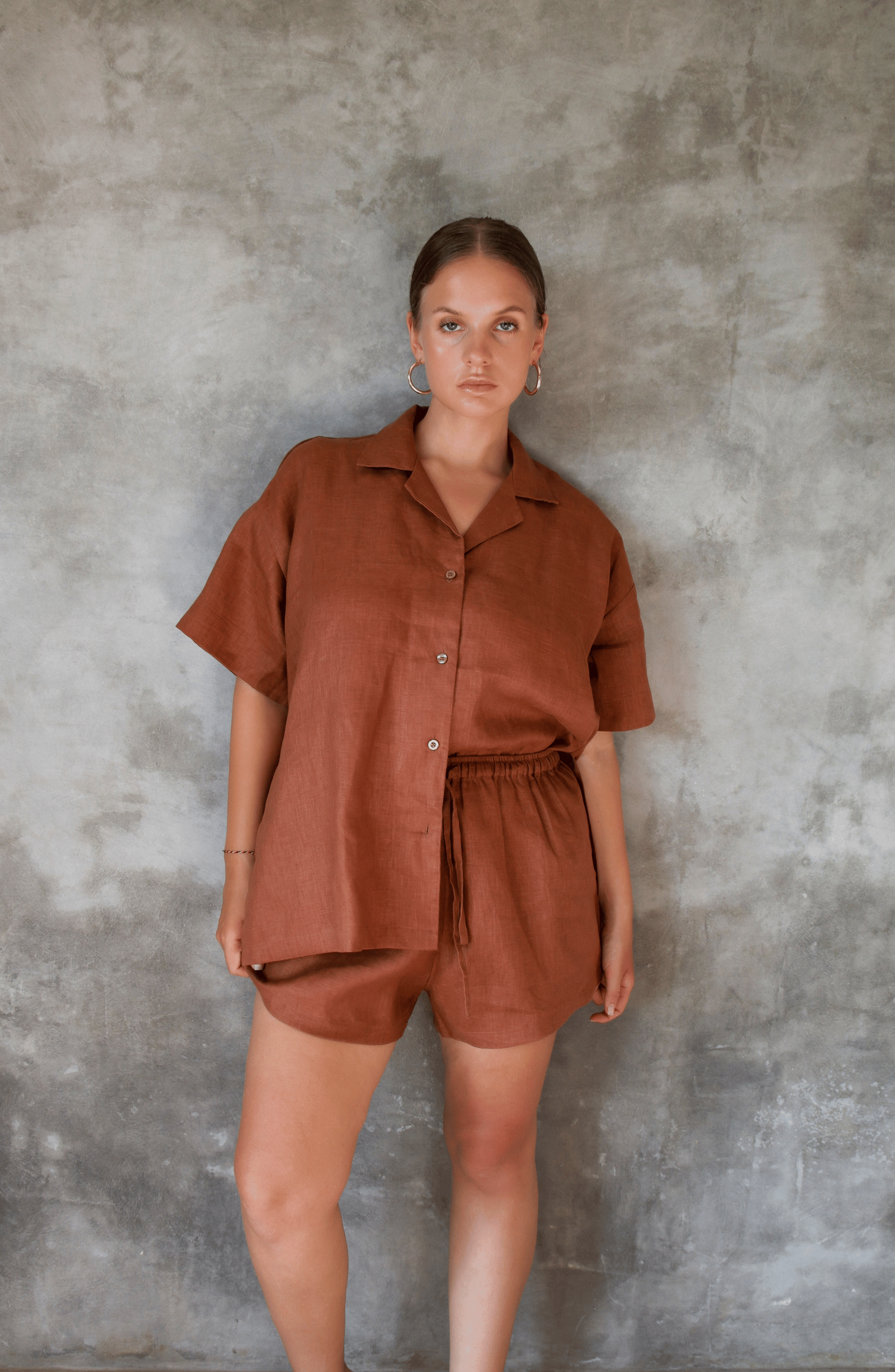 Model poses for photo in Pinksalt Swimwear klum espresso brown Linen shirt and shorts set. This is a front view, with one side tucked in, hands down by her side.
