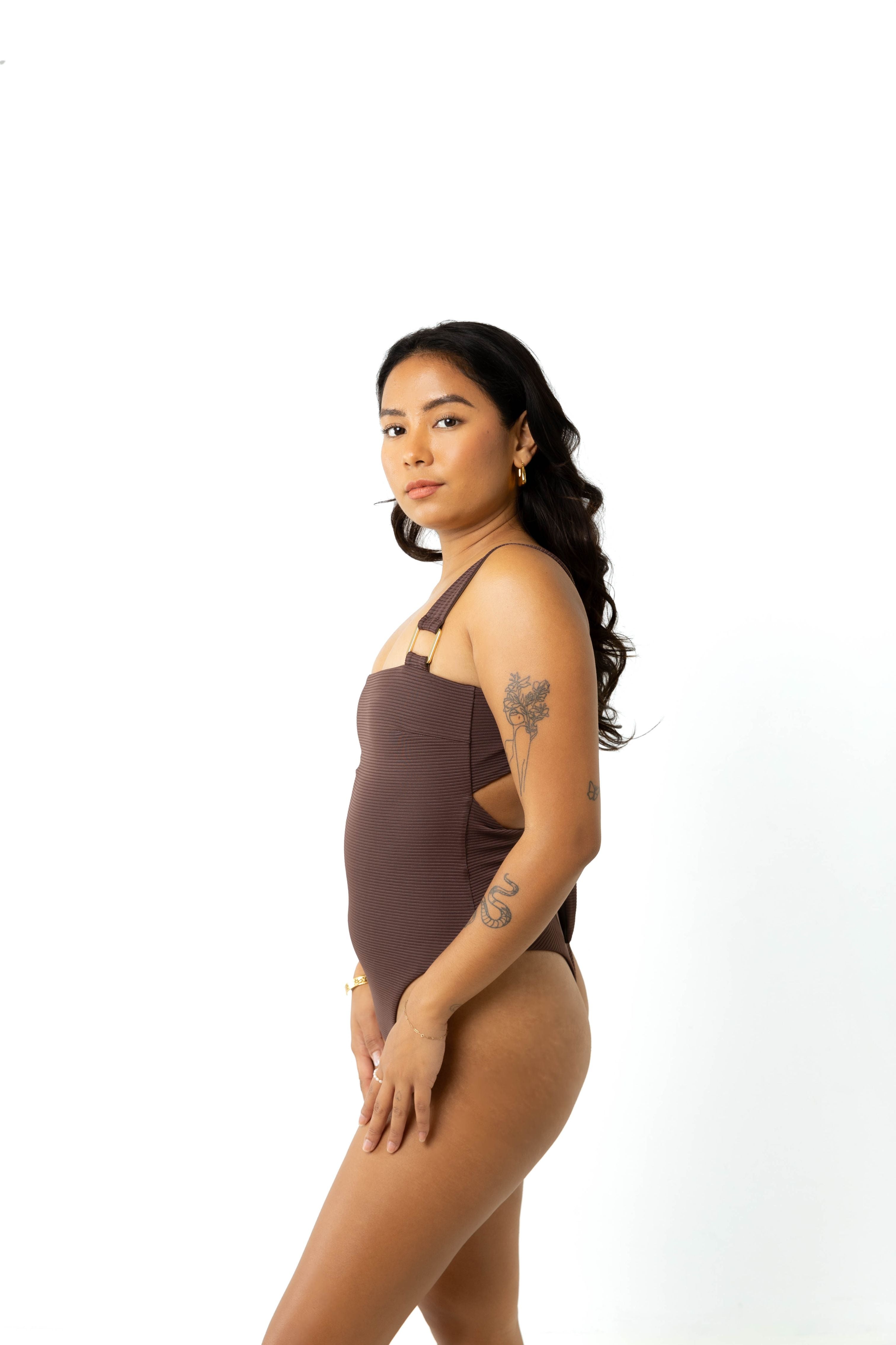 Size 6 in studio wearing cheeky moss chocolate brown eco ribbed one piece swimsuit with adjustable straps, view from the side