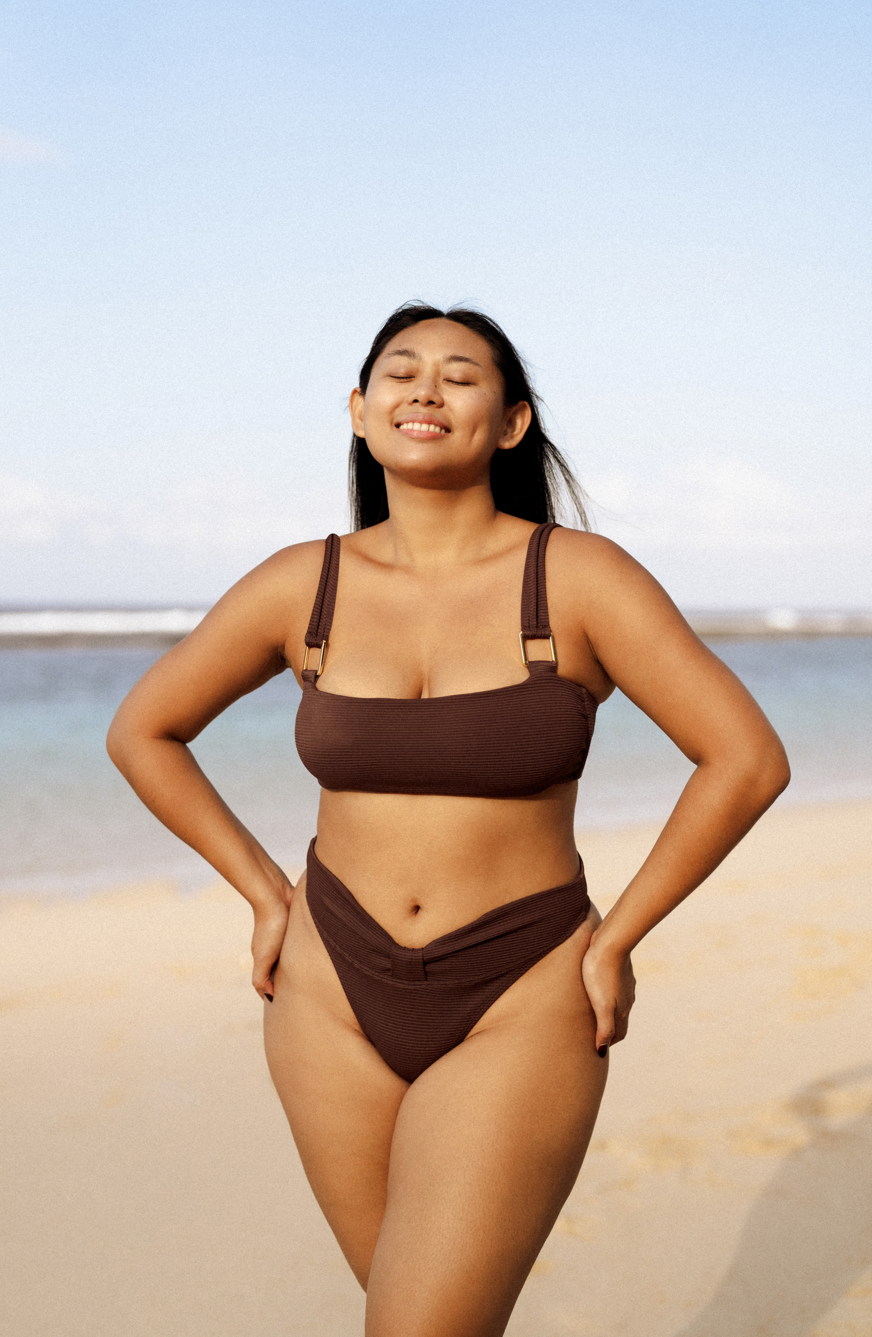 Woman in a brown bikini standing on a beach with a clear sky.