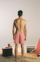 A man stands with his back to the camera wearing red and white gingham checkered swim trunks, set against a minimalist studio background with vintage props.
