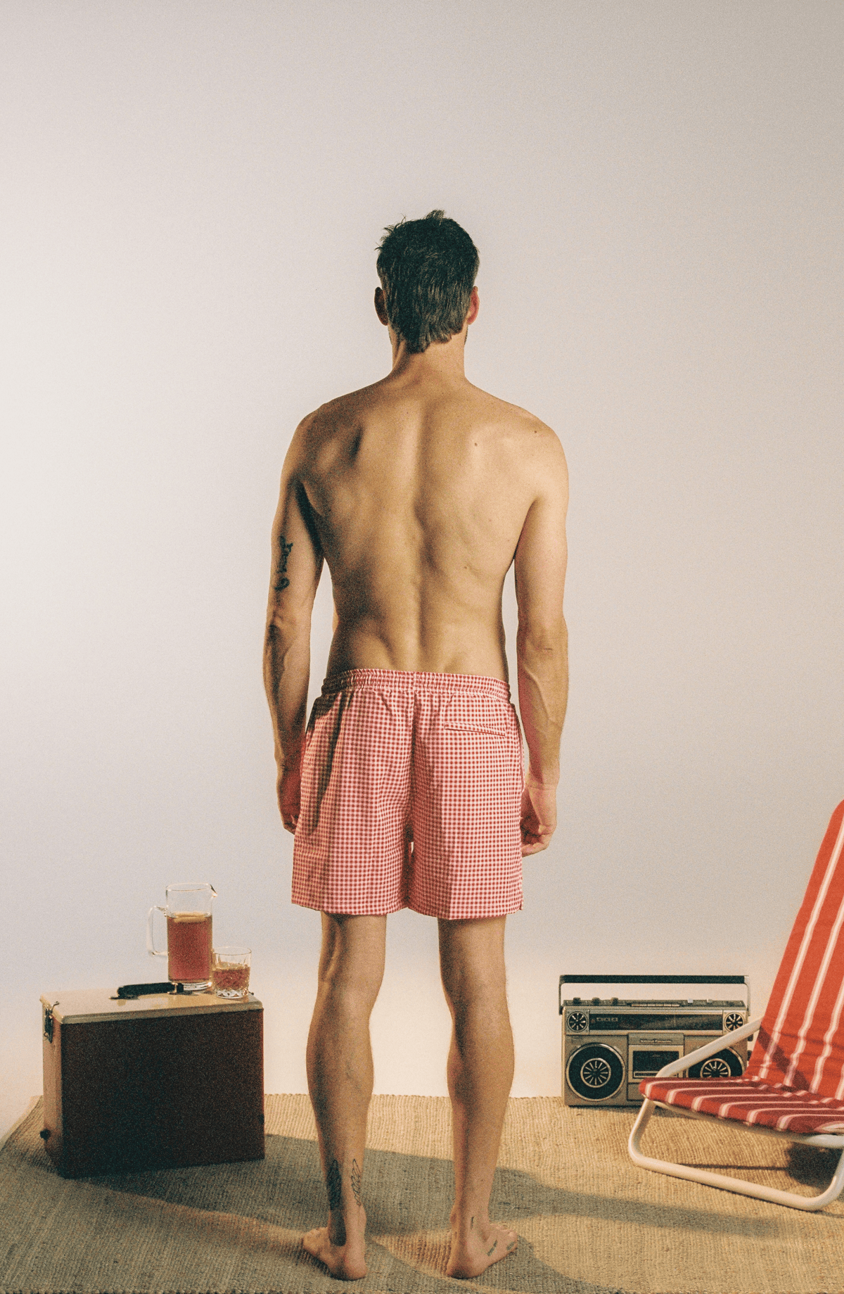 A man stands with his back to the camera wearing red and white gingham checkered swim trunks, set against a minimalist studio background with vintage props.