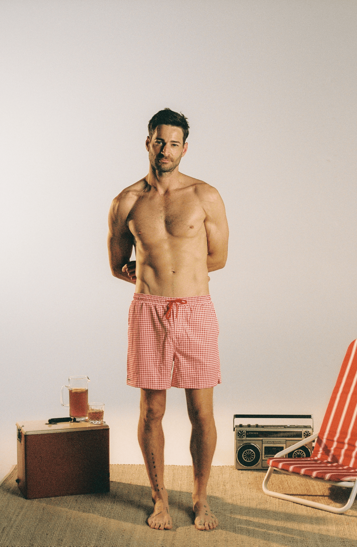A shirtless man stands barefoot in a studio setting wearing red and white gingham check swim trunks. A vintage suitcase with a drink, a retro boombox, and part of a striped beach chair are placed on the floor around him against a plain light background.
