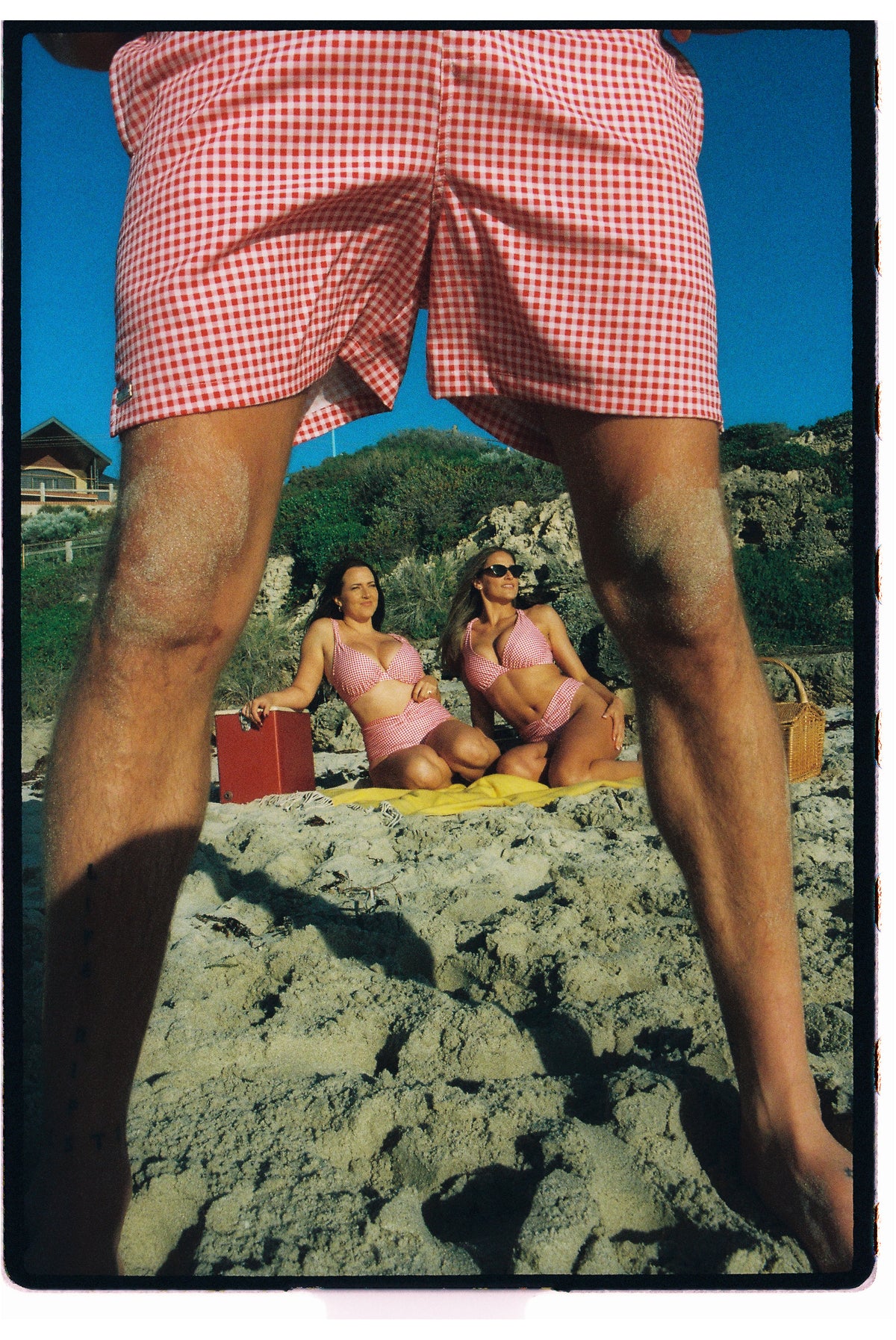 A person wearing red and white gingham check boardshorts stands on a sandy beach, framing two women in red and white gingham bikinis sitting on a yellow towel in the background under a blue sky.