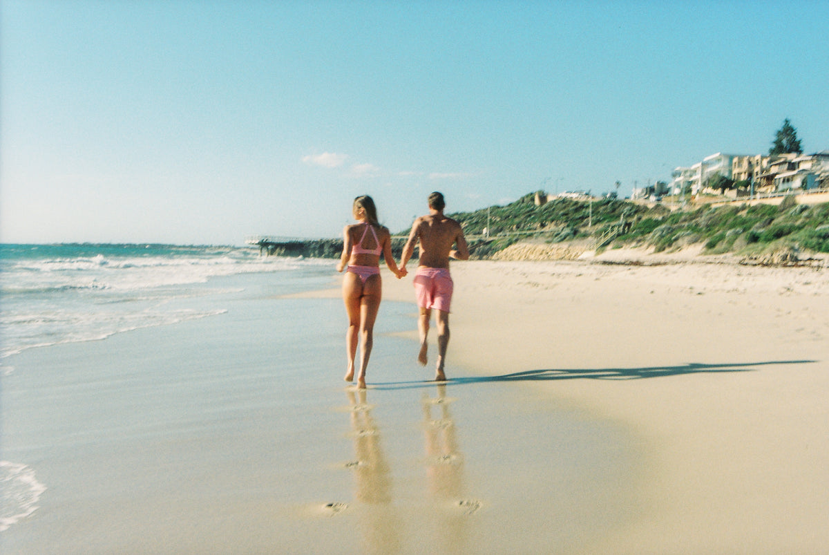Couple wearing matching Pinksalt Cherry Gingham swimwear and Arthur boardshorts running on the beach