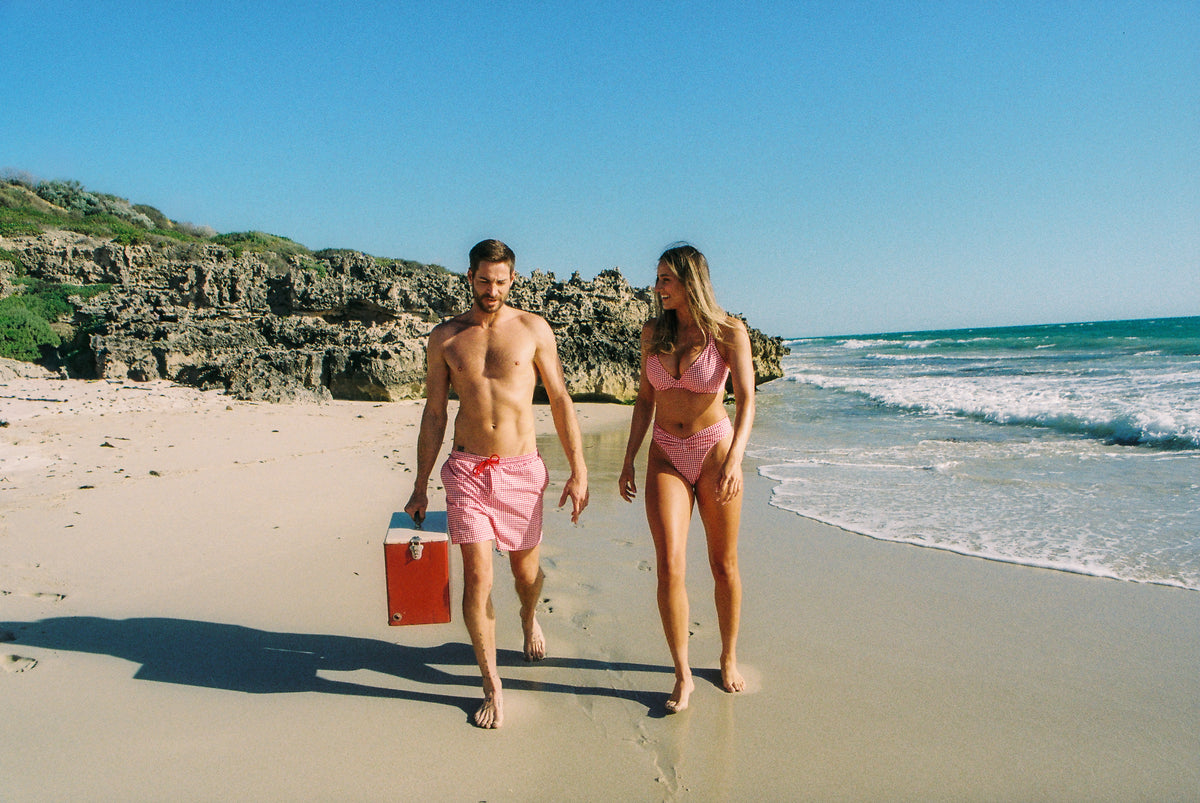 Couple wearing matching Pinksalt Cherry Gingham swimwear and Arthur boardshorts walking on the beach with esky in hand