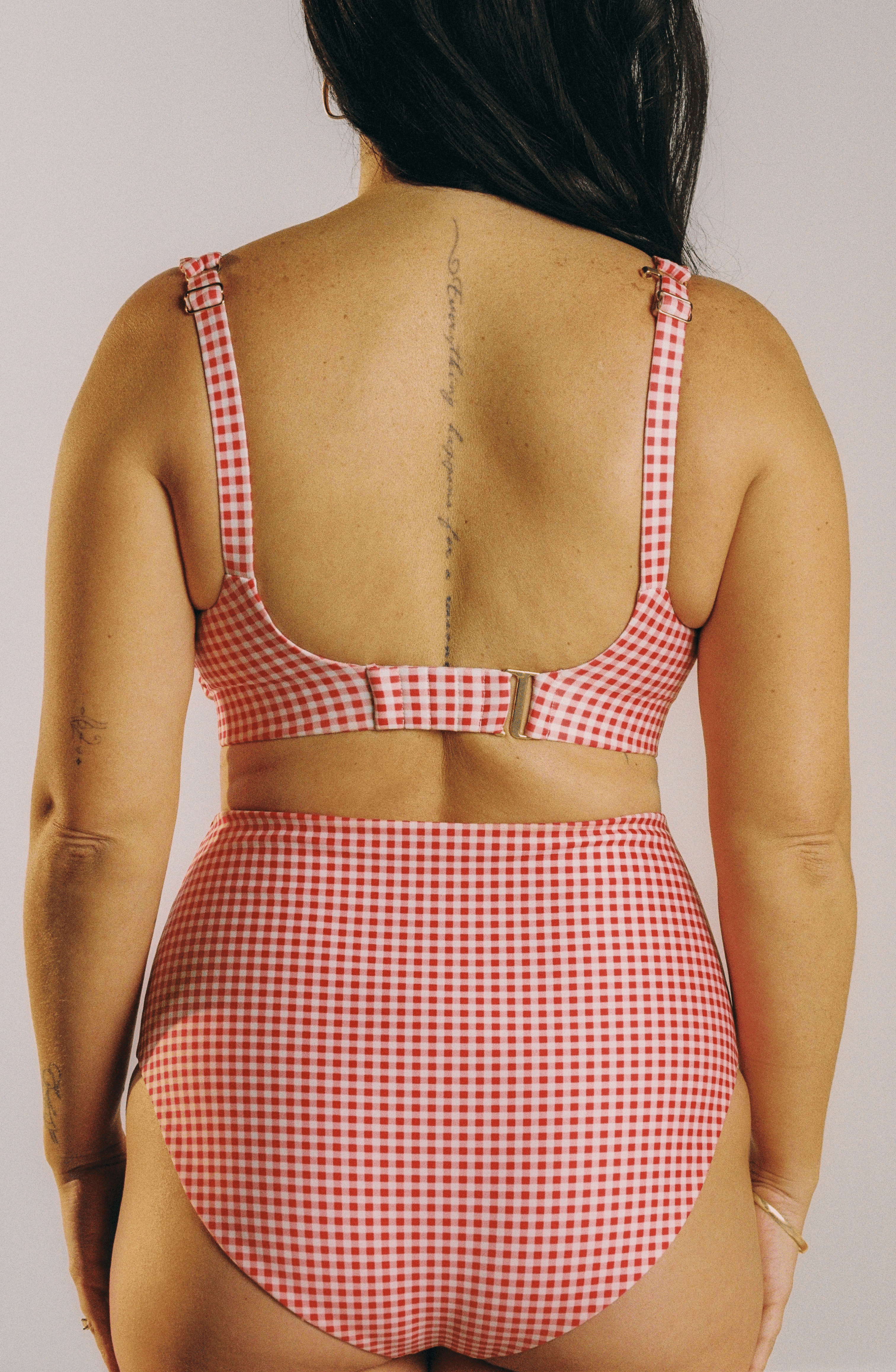 A woman shown from the back wearing a red and white gingham two-piece swimsuit with a cutout waist and adjustable straps, against a plain light white studio background.