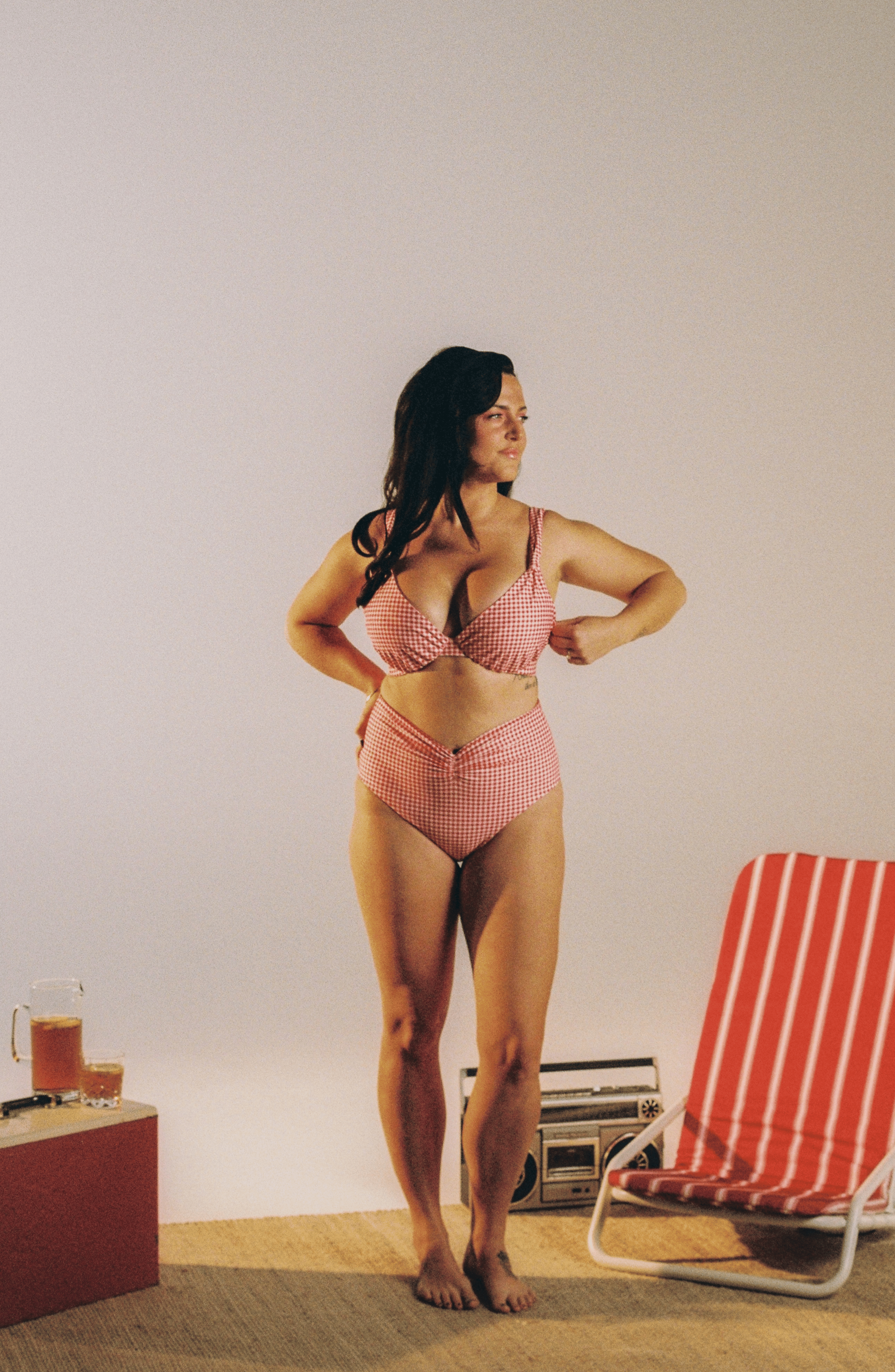A woman stands barefoot indoors wearing a red and white gingham two-piece bikini. A red-and-white striped lounge chair and a small table with drinks are visible against a plain light wall.