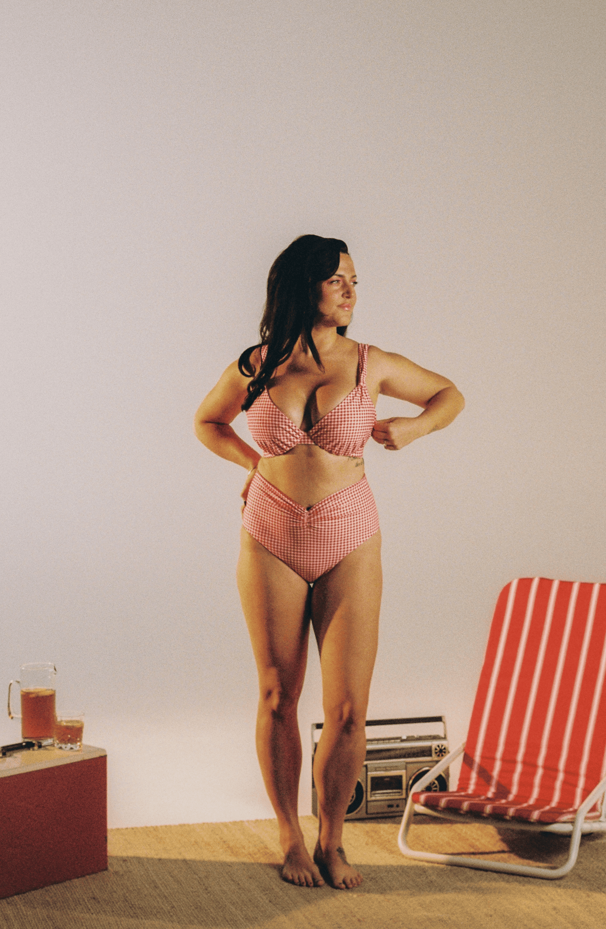 A woman stands barefoot indoors wearing a red and white gingham two-piece bikini. A red-and-white striped lounge chair and a small table with drinks are visible against a plain light wall.
