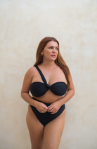 swimwear model standing against wall facing camera wearing Pinksalt Swimwear - a black one shoulder bikini top and black high waisted bikini bottoms 