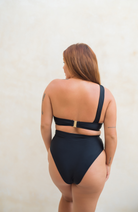woman with long red hair facing her back to the camera wearing her Pinksalt Swimwear. The woman is wearing a black one shoulder sliding bandeau bikini top with gold clip  and black high waisted bottoms.