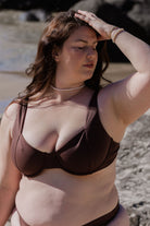 A model wearing a chocolate brown underwire bikini top, with the focus on the product and the surrounding beach environment.