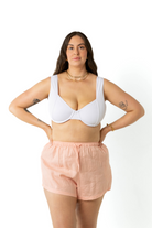 model in a photography studio facing camera, wearing a white ribbed underwire bikini top with peach colour linen shorts with her hands placed on her hips.