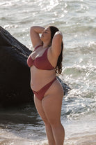 A woman wearing a high-cut leg spice pink bikini bottom standing near a rock at the beach.