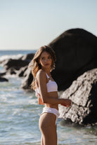 A woman standing by the ocean wearing high-cut white bikini bottoms and a matching top, with her hands held in frot of her looking back.